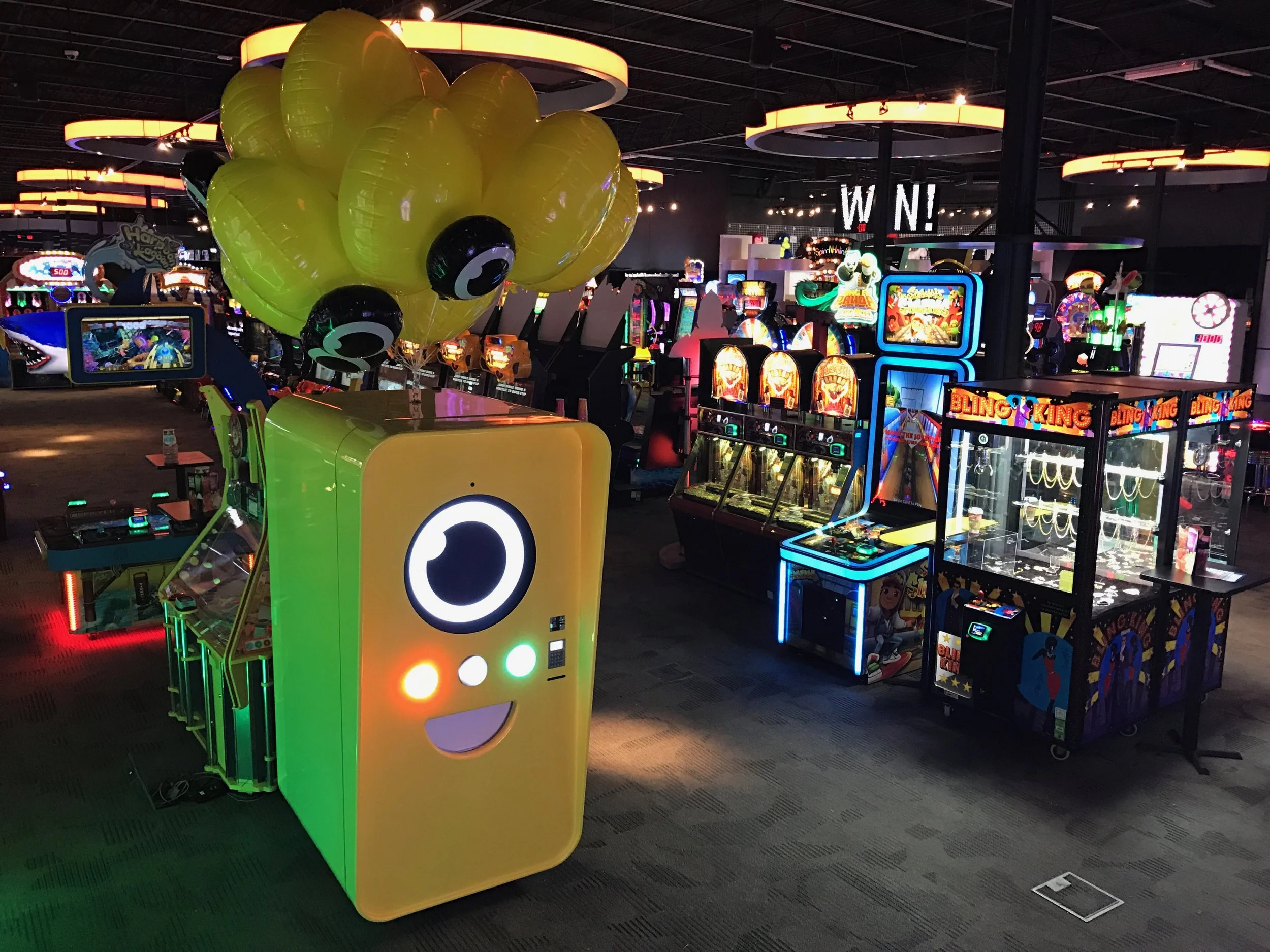 Yellow Snapbot vending machine with eyeball balloons on a Dave and Buster's arcade floor surrounded by game cabinets
